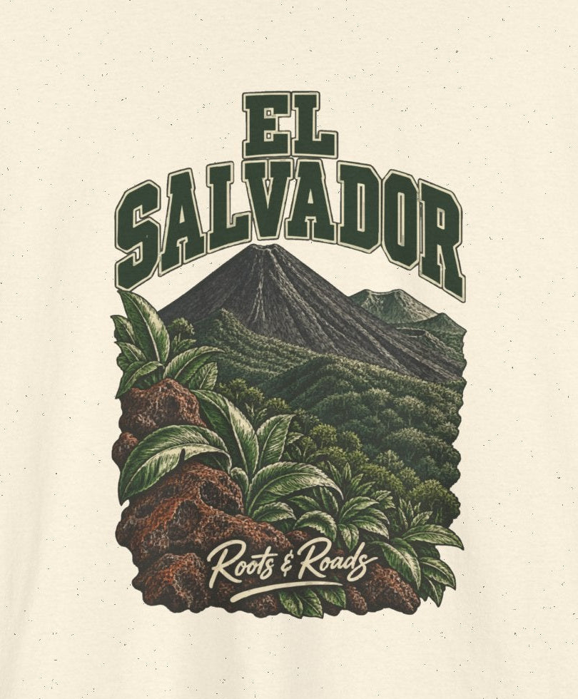 Print detail cropped from the Natural El Salvador supplier T-shirt mockup, showing green two-line EL SALVADOR lettering, a forested volcanic cone, broad leaves and warm lava rocks and Roots & Roads script.