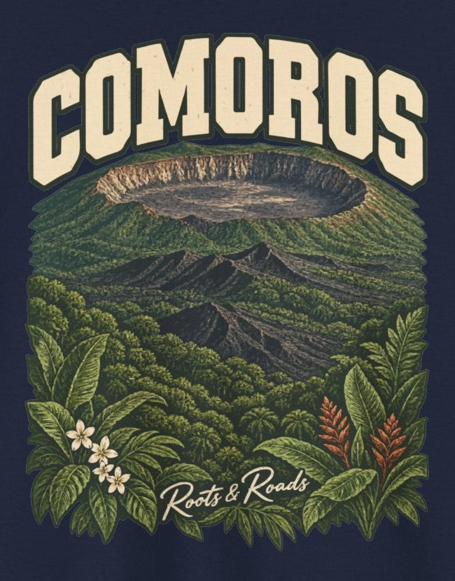 Print detail cropped from the Navy Comoros supplier T-shirt mockup, showing cream COMOROS lettering above a volcanic caldera, green forest and flowering foliage and Roots & Roads script.