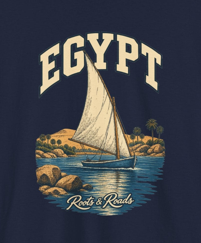 Print detail cropped from the Navy Egypt supplier T-shirt mockup, showing cream EGYPT lettering, a triangular-sailed boat, blue river water, palms and warm rocks and Roots & Roads script.