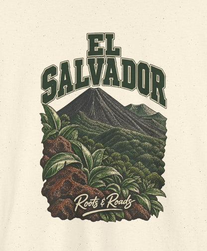 Print detail cropped from the Natural El Salvador supplier T-shirt mockup, showing green two-line EL SALVADOR lettering, a forested volcanic cone, broad leaves and warm lava rocks and Roots & Roads script.