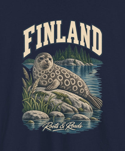 Print detail cropped from the Navy Finland supplier T-shirt mockup, showing cream FINLAND lettering, a ringed seal, lake water, reeds and wooded islands and Roots & Roads script.