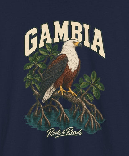 Print detail cropped from the Navy Gambia supplier T-shirt mockup, showing cream GAMBIA lettering, a white-headed fish eagle, mangrove roots and still water and Roots & Roads script.