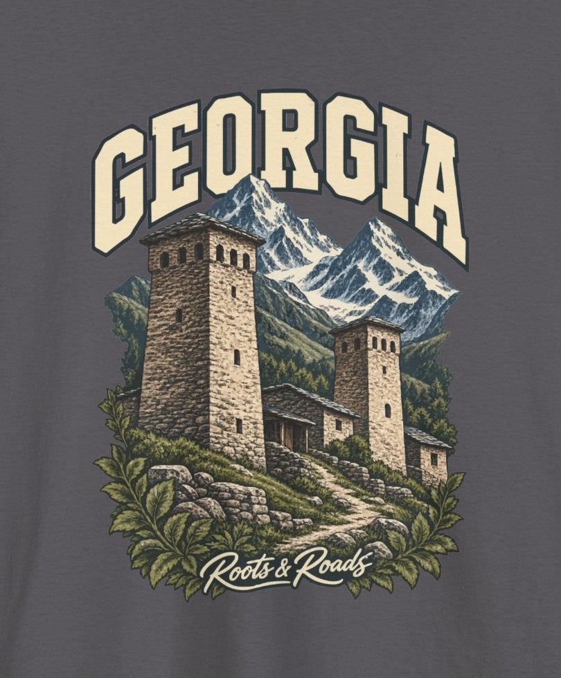 Print detail cropped from the Charcoal Georgia supplier T-shirt mockup, showing cream GEORGIA lettering, stone towers, snowy mountains, clustered buildings and a winding path and Roots & Roads script.